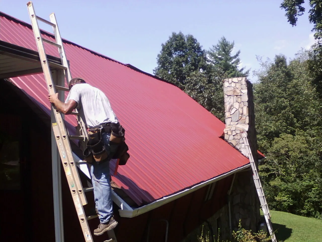 Roofer on a ladder beside a red metal roof and stone chimney — Soffit & Fascia Repair by Roofing Brothers in Union Bridge